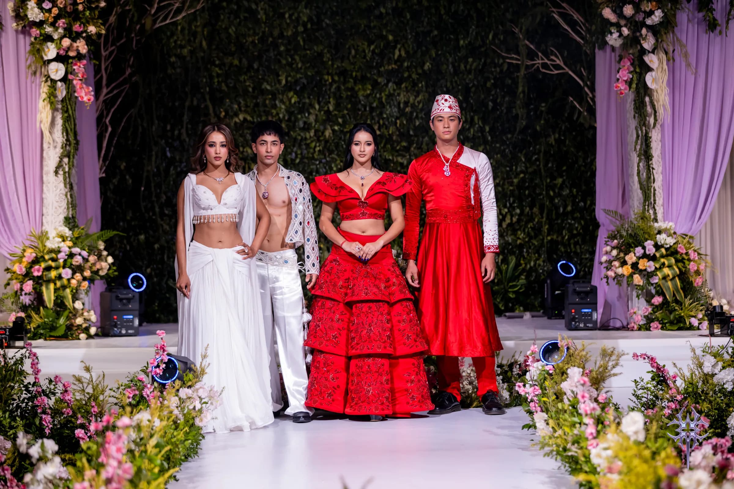 Four models on the Asteria runway — white drapes and a mirror-work jacket beside red embroidered couture — on a flower-lined aisle